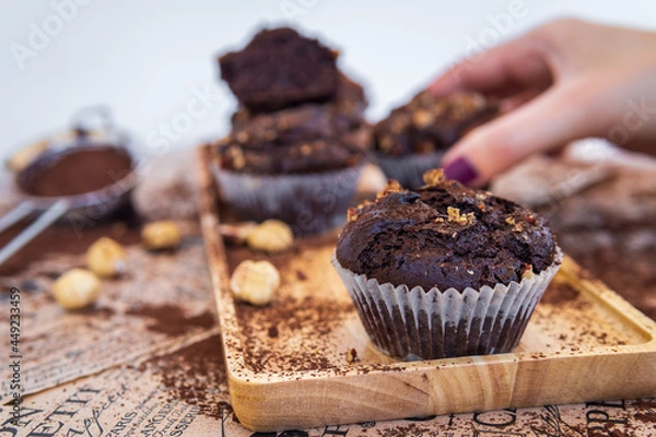 Fototapeta Healthy gluten free chocolate muffin in a baking paper cup with pecan nuts and cocoa powder. Homemade, freshly baked delicious cupcakes in the background and woman's hand takng one. Nutrition concept