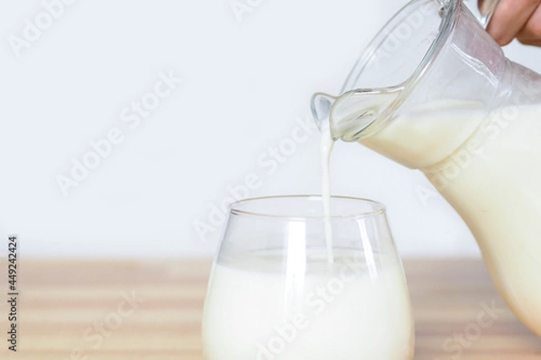 Obraz hand pours milk from a jug into a glass on a wooden table on a gray background..