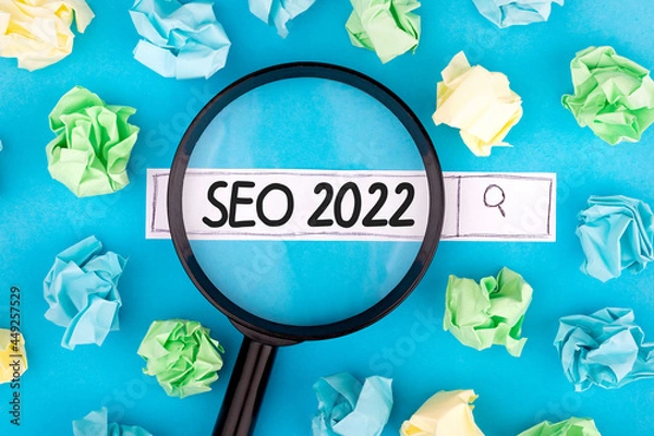 Fototapeta Concept of searching. Text SEO 2022 with magnifier and pieces of paper on blue background.