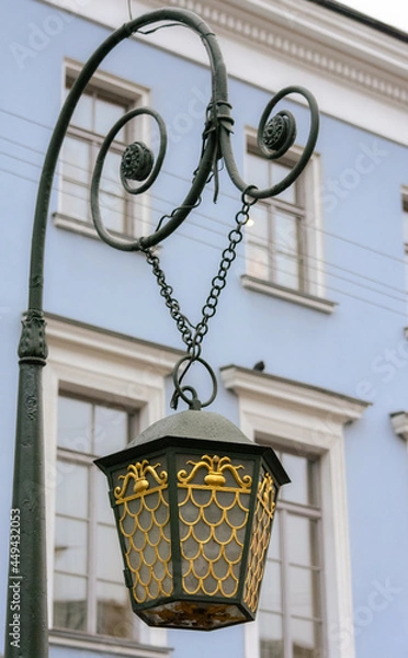 Fototapeta Fragment of an old street lamp hanging on a chain against the background of a blue building