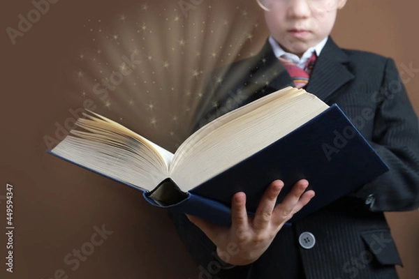 Obraz smart child 7-8 years old, kid in black school suit stands and reads thick tome, open paper book with magic glow and question marks, concept young child prodigy, back to school