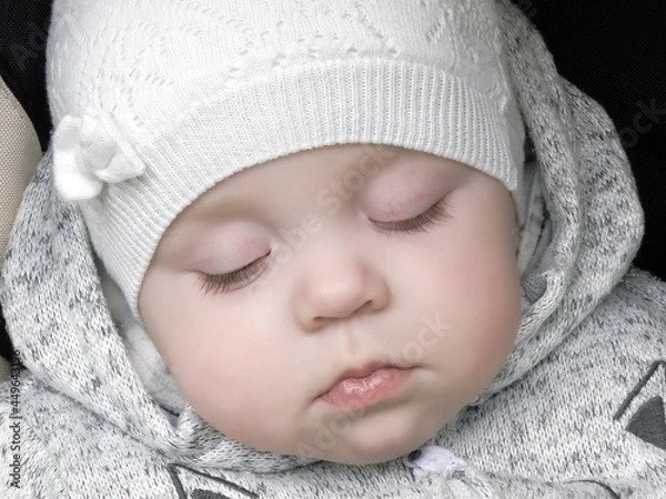 Fototapeta Portrait of a sleeping little baby wearing a creamy cap. Soft alabaster skin. Delicate baby dressed  for a walk. Calm healthy sleep outside. 