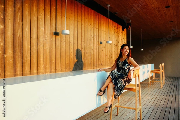 Fototapeta Asian woman sitting on high chair at the bar counter and watching the sunset