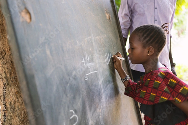 Obraz Concentrated little black girl writing numbers on a blackboard during open air classes in a rural community in West Africa