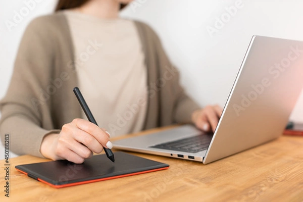 Fototapeta Unrecognizable girl in beige t-shirt and grey jacket sitting at wooden table opposite laptop and drawing picture on graphic tablet. Designer remotely work from home. Soft focus on hand with pencil.