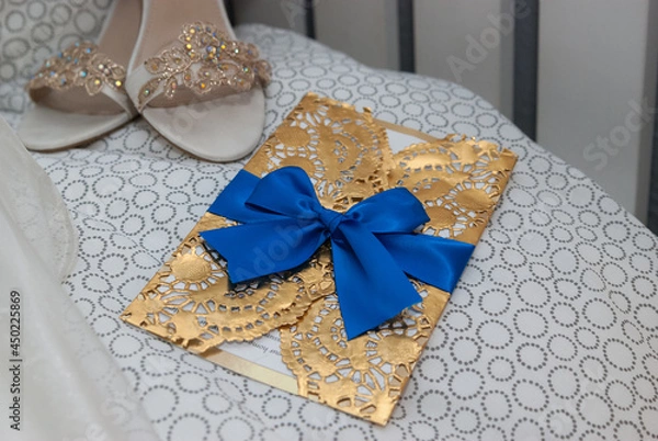 Fototapeta Invitation in a golden envelope decorated with a blue ribbon next to a beautiful pair of high heels decorated with lace.