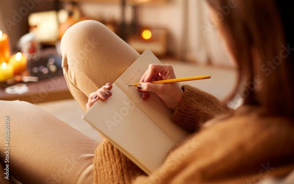Fototapeta halloween, holidays and leisure concept - close up of young woman with pencil writing to diary and resting her feet on table at cozy home