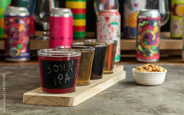 Fototapeta Craft beer tasting, sour fruit IPA, lager, stout, and pale ale. Bar with different cans in the blurred background. Wooden board with flight set. 