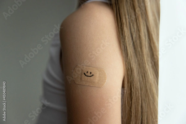 Fototapeta Little girl with long brown hair in white tank top got her vaccine, little's girl arm with plaster with drawn smile after vaccine