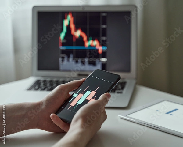 Obraz Millennial unknown male analysing graphs and data on laptop and smartphone at office. Online trader and shareholder. Faceless young businessman buying stocks. Trader or investor working at home