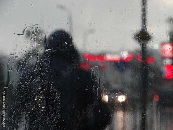 Fototapeta Wet glass surface after rain, soft focus. Night city, street and cars lighting blurry. Water drops. Nostalgic. Petrol station, human figure, signboard defocused by rain drops on a window glass.