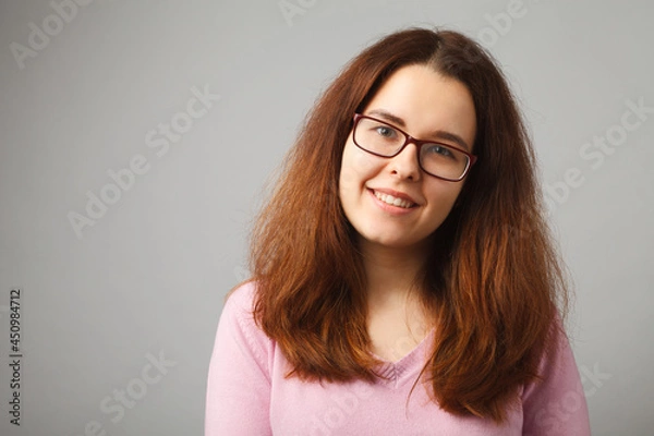 Fototapeta Redhead long-haired girl with glasses smiles, looking at the camera. Studio girl portrait on a solid light background