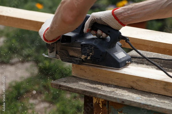 Obraz A carpenter planks wood with an electric plane. Woodworking carpentry. Close-up. Man in the garden sanding wooden planks. DIY home improvement, restoration, carpentry concept. Sawdust flies