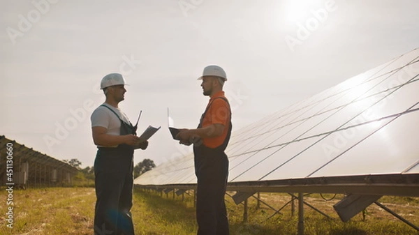 Fototapeta Team of industrial colleagues using tablet monitoring rows of photovoltaic solar panels at sunset. Business cooperation. Solar park. Alternative energy concept. Construction solar power plant.