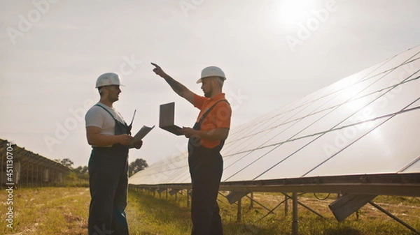 Fototapeta Team of industrial colleagues using tablet monitoring rows of photovoltaic solar panels at sunset. Business cooperation. Solar park. Alternative energy concept. Construction solar power plant.