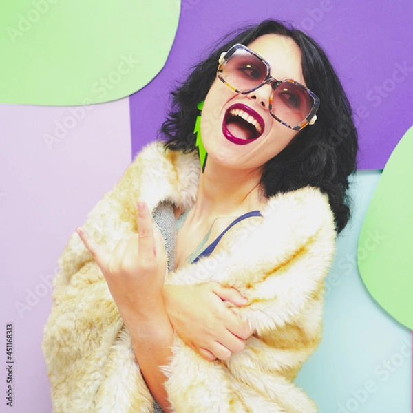Obraz Beautiful woman in dark glasses, with dark red lipstick, in a white faux fur coat and earrings on the background of pop art smiles and shows a rocker sign