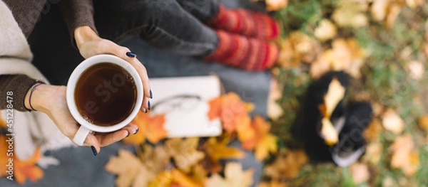 Fototapeta Woman taking picnic in autumn forest. In hands she hold white cup with black tea. On the plaid lying book, glasses and orange maple leaves. Autumn concept. Selective focus on a mug. Copy space.