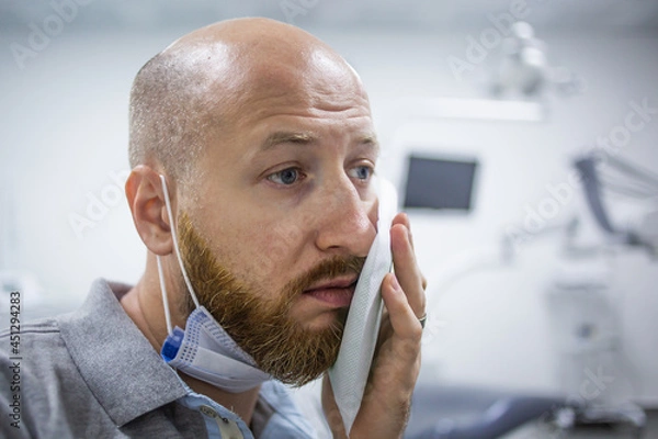Fototapeta Unhappy and scared caucasian male in a dental clinic waiting for a check up and holding a cooling element at his cheek because of a toothache