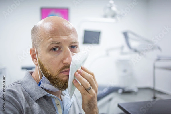 Fototapeta Unhappy and scared caucasian male in a dental clinic waiting for a check up and holding a cooling element at his cheek because of a toothache