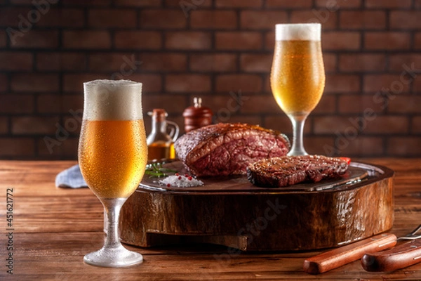Fototapeta Two sweaty cold tulipa glasses of beer with grilled sliced cap rump steak on wooden cutting board (Brazilian picanha).