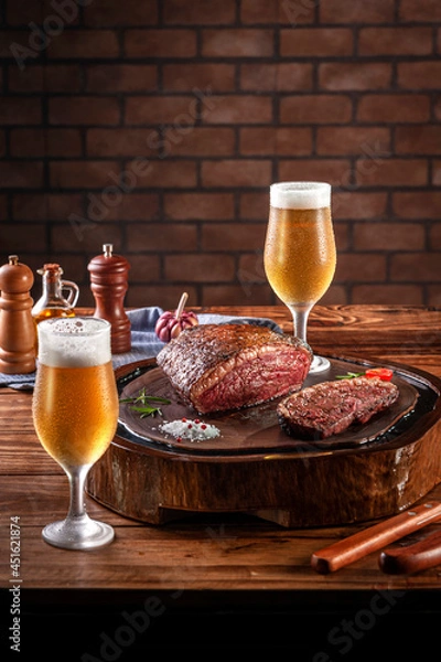 Fototapeta Grilled cap rump steak on wooden cutting board with two sweaty cold tulipa glasses of beer. Wooden table and bricks wall background (Brazilian picanha).