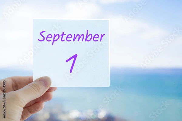 Fototapeta 1st September. First day of autumn. Knowledge day. Hand holding sticker with text September 1 on the blurred background of the sea and sky. Copy space for text. Month in calendar concept