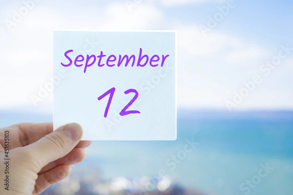 Fototapeta 12th September. Hand holding sticker with text September 12 on the blurred background of the sea and sky. Copy space for text. Month in calendar concept