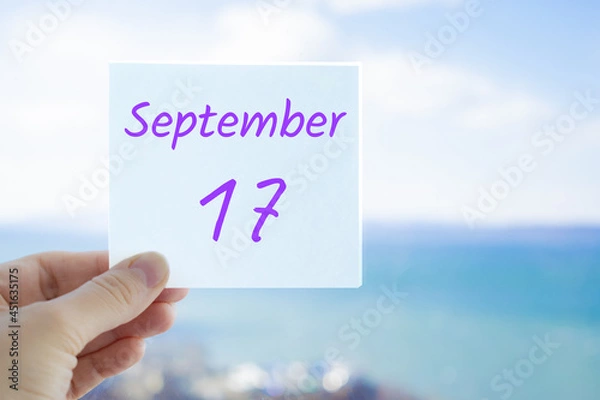 Fototapeta 17th September. Hand holding sticker with text September 17 on the blurred background of the sea and sky. Copy space for text. Month in calendar concept