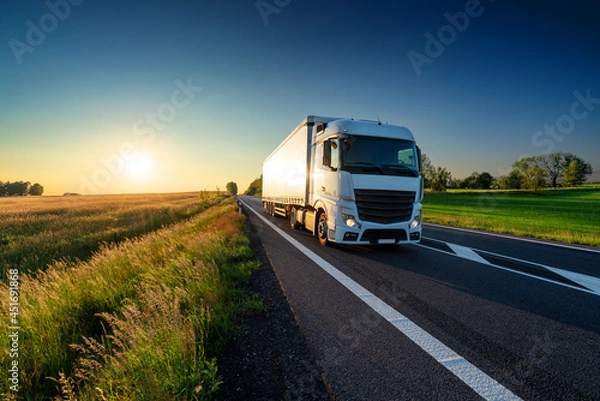 Obraz White truck driving on the asphalt road in rural landscape at sunset