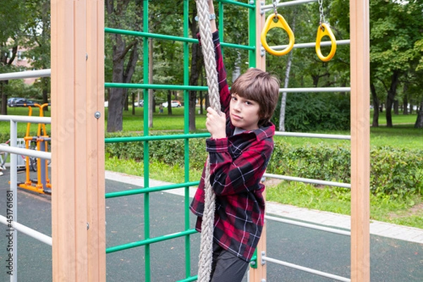 Fototapeta Boy in red plaid shirt playing on crossbars in playground in summer on background of green trees