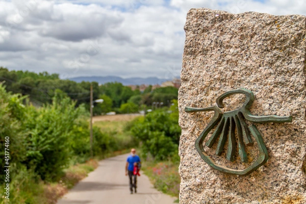 Fototapeta Pilgrim out of focus walks along the Camino de Santiago (Sant James Way) carrying a backpack next to a column on focus with the shell symbol of the Camino Santiago de Compostela