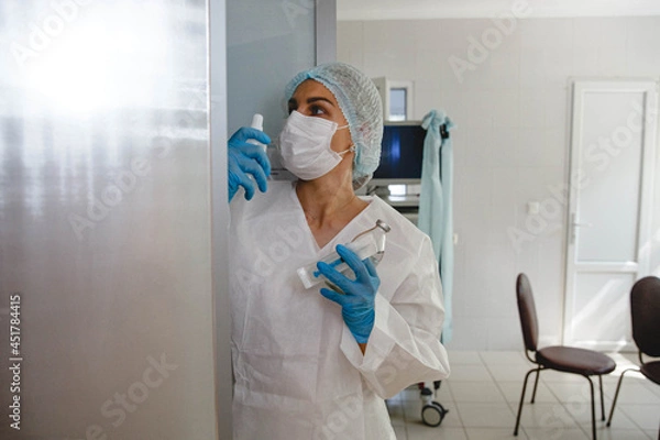 Fototapeta A young nurse in a protective suit, cap, mask and gloves takes medications from a locker in the medical office