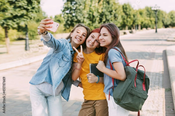 Fototapeta Three charming friends take selfies with their phone on the way to school. The concept of friendship. Education, training, back to school