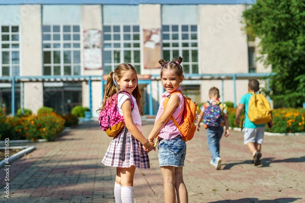 Fototapeta Two girls, elementary school students with backpacks, walking down street Happy children are returning to school. beginning of school year. Children in full growth, happily went to school. rear view