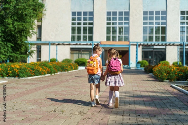 Fototapeta Two girls, elementary school students with backpacks, walking down street Happy children are returning to school. beginning of school year. Children in full growth, happily went to school. rear view