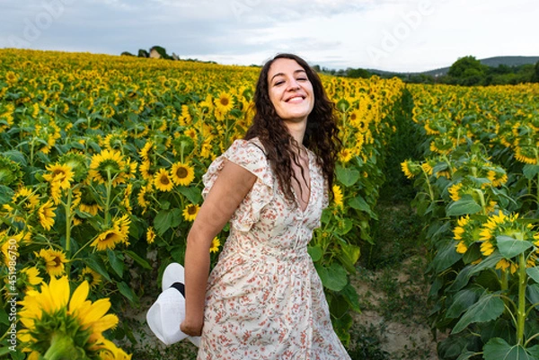 Fototapeta Smiling young woman walking in the middle of a field of sunflowers