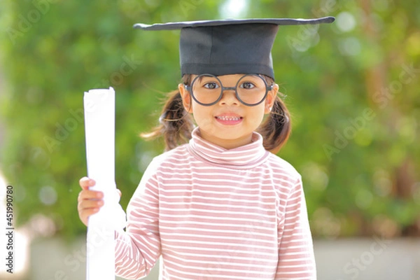 Fototapeta A little girl asia looks happy while celebrating his graduation and holding a diploma and graduation cap  which increases the development and enhances outside the classroom learning skills concept.