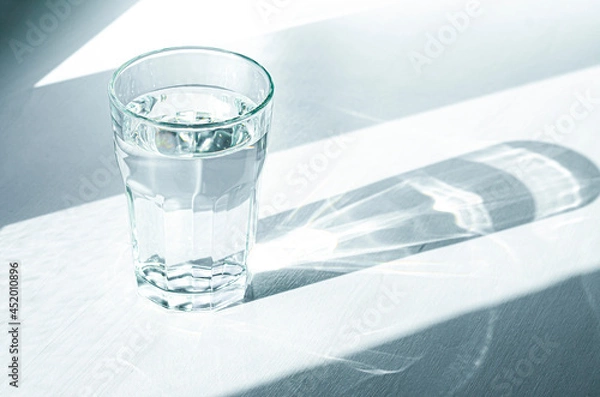 Fototapeta A glass with clean clear water and sharp shadows stands on a white table