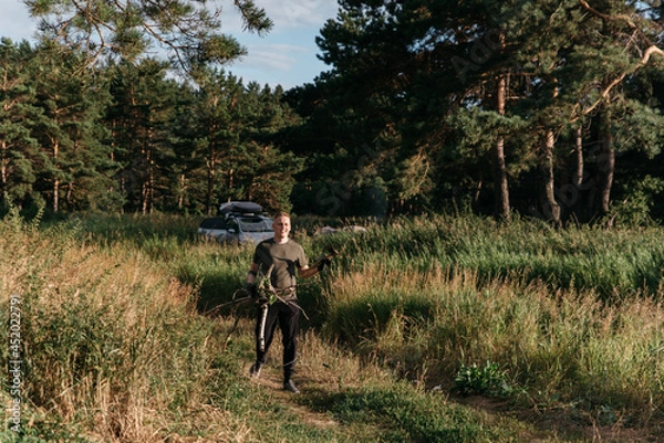 Fototapeta Photo of a Blond Man on a Summer Camping Activity with firewood for bonfire. Travel location of popular tourist outdoor. View of beautiful Forest Landscape. Vacation in the lake in a camp with a tent