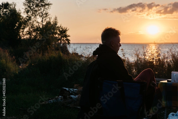 Fototapeta Photo of Man in a Summer Camping Activity. Travel location of popular tourist outdoor. View of beautiful Sea Landscape. Vacation by the lake in a camp with a bonfire and tent