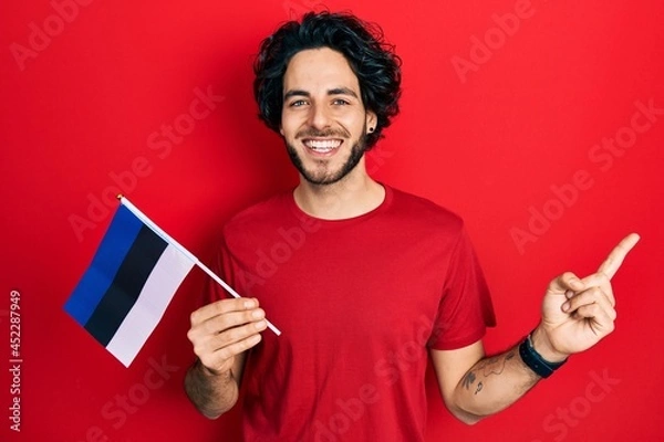 Obraz Handsome hispanic man holding estonia flag smiling happy pointing with hand and finger to the side