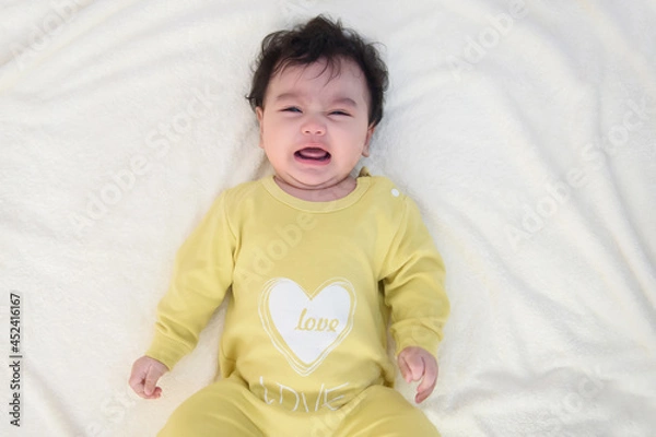 Obraz Top view of a cute little Asian baby girl wearing yellow dress, lying on the bed, sad and crying