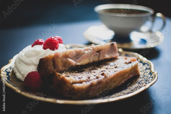 Fototapeta Delicious sweet baked homemade banana bread with nuts, raisins, whipped cream and raspberries on small dessert ornamental plate. A cup of tea in the background.  Vintage design. Selective focus.