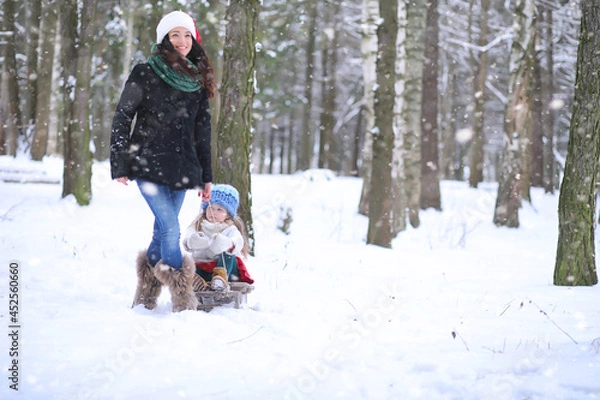 Obraz A winter fairy tale, a young mother and her daughter ride a sled in the forest. A girl on a sled with gifts on the eve of the new year in the park. 