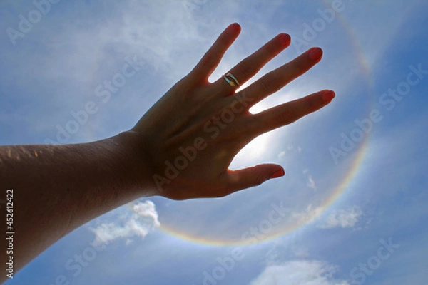 Obraz Woman's hand with ring and painted nails covering the sun in a solar halo effect, on a celestial sky.