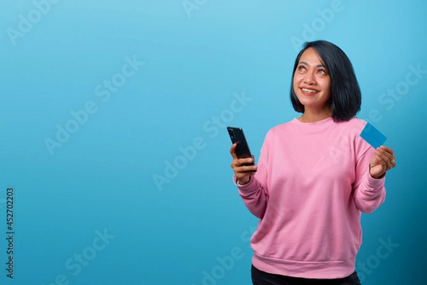 Obraz Portrait of beautiful smiling asian woman using mobile phone and holding credit card and looking up empty space