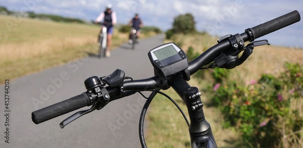 Fototapeta Konzept: Pause am Rand eines Fahrradweges. Nahaufnahme eines Lenker eines E-Bikes im Vordergrund, Fahrradfahrer, Weg und Natur im Hintergrund, Querformat, selektiver Fokus