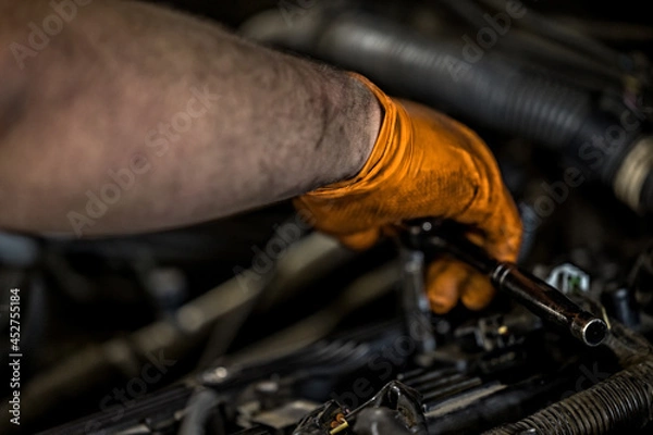 Fototapeta A man wearing an orange glove fixing and doing maintenace on a car engine in an auto repair shop