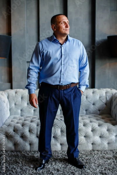 Obraz Handsome stylish man in a blue shirt at home, standing near a gray sofa. Full-length business portrait