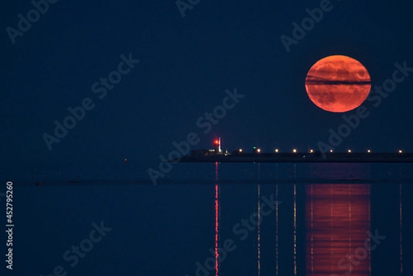 Fototapeta Beautiful view of rare summer full blue sturgeon Moon rising over Irish sky between Dun Laoghaire lighthouses on August 23, 2021 seen from Sandymount Beach, Dublin, Ireland. Huge Moon